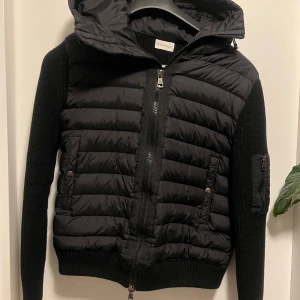 Svart jacka från Moncler - Säljer en snygg svart jacka från Moncler med quiltad design och stickade ärmar. Jackan har en huva och dragkedja framtill samt en broderad logga på ärmen. Perfekt för kyligare dagar! Denna cardigan är i storlek M men passar även S, hör av er vid minsta fundering.