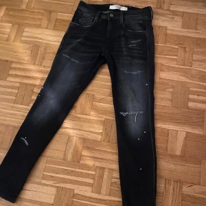 Svarta jeans från Replay - Jeansen sitter slim och som 29/30, Snygga svarta jeans från Replay med en cool slitningseffekt. De har en klassisk femficksdesign och en knappgylf. Perfekta för en stilren look.
