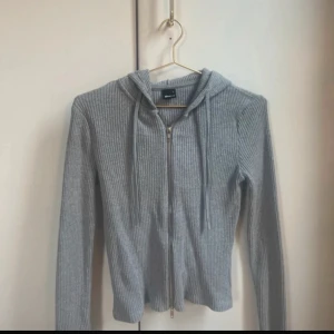 Grå ribbad hoodie - Säljer en stilren grå ribbad hoodie med dragkedja och huva. Tröjan har långa ärmar och är perfekt för en avslappnad look. Passar bra till både jeans och mjukisbyxor.