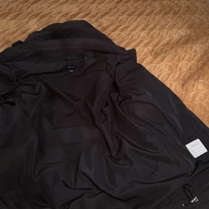 Svart dunjacka från GANT - Säljer en svart dunjacka från GANT i storlek 176 cm. Jackan har en dragkedja framtill och praktiska fickor med knappar. Perfekt för kyliga dagar med sin vadderade design och huva.