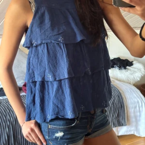 Blå ärmlös topp med volanger - Snygg blå ärmlös topp med flera lager av volanger. Perfekt för en somrig look. Toppen har en luftig passform och är tillverkad i ett lätt material.