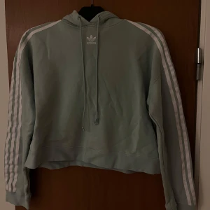Turkos hoodie från Adidas - Säljer en snygg croppad hoodie från Adidas med de klassiska tre ränderna längs ärmarna. Tröjan har en justerbar huva med dragsko och en avslappnad kortare passform. Nypris för denna 529 på aboutyou. Pris går att diskutera