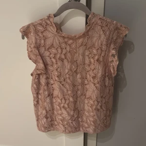Rosa spetsblus - Säljer en söt rosa spetsblus med volangdetaljer. Blusen är ärmlös och har ett vackert blommönster. Perfekt för en somrig look!