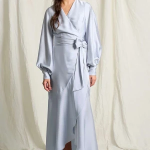 Ljusblå omlottklänning från Joelle - Elegant ljusblå långklänning i satin med omlott-design och v-ringning. Klänningen har långa ärmar med puffiga manschetter och en knytning i midjan som ger en fin siluett. Perfekt för speciella tillfällen.Ny med lappar kvar.Storlek 44. Butikspris 1499kr