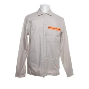 Beige overshirt från Hugo Boss - Snygg beige overshirt från Hugo Boss i 100% bomull. Den har en avslappnad passform med långa ärmar och en cool orange detalj på bröstfickan. Perfekt för en stilren look.