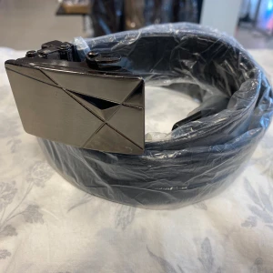 Svart bälte med geometriskt spänne - Snyggt svart bälte med ett geometriskt mönstrat spänne i metall. Perfekt för att ge en stilren touch till din outfit. Bältet är tillverkat i skinn och har en modern design.