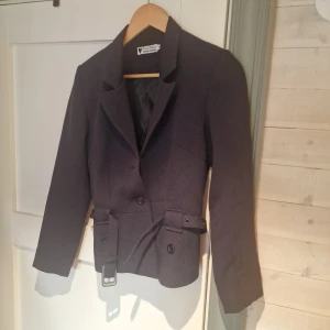 Mörkblå kavaj från Daisy Dapper - Snygg mörklila kavaj från Daisy Dapper Retro Clothing. Kavajen har en klassisk design med knappar och ett bälte i midjan. Bliderna gör den inte rättvis!! 