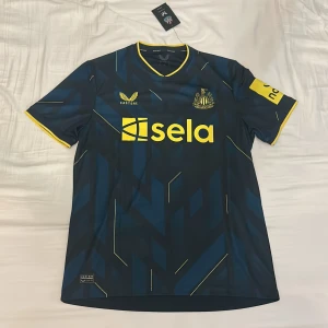 Newcastle United tredje-tröja 23/24 - Newcastle Uniteds tredje-tröja från säsongen 2023/24. Tröjan är helt ny och har lapparna kvar. Den är köpt på classicfootballshirts.com, och är alltså helt äkta.