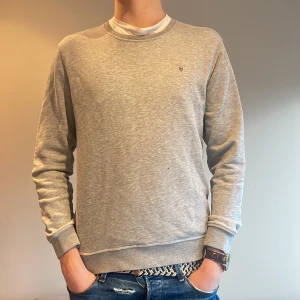 Grå sweatshirt från Gant - Säljer en stilren grå sweatshirt från Gant med rund halsringning och broderad logotyp på bröstet. Tröjan har långa ärmar och ribbade muddar vid ärmslut och nederkant. Perfekt för en avslappnad look. Lite mindre i storleken. En fläck på högra nedre bröstet men går säkert bort i tvätten.