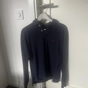 Mörkblå pikétröja från Ralph Lauren - Säljer en stilren mörkblå pikétröja från Ralph Lauren med klassisk krage och knappar. Tröjan har långa ärmar och en liten röd logotyp på bröstet. Använda ett par gånger