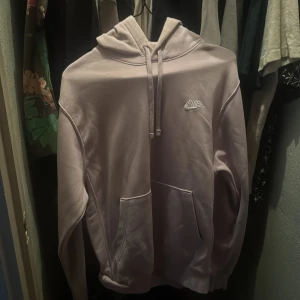 Ljusrosa hoodie från Nike - Säljer en stilren ljusrosa hoodie från Nike med broderad logga på bröstet. Tröjan har en klassisk design med känguruficka och justerbar huva. Perfekt för en avslappnad stil.