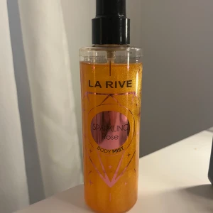 La Rive Sparkling Rose Body Mist - Fräsch och glittrande body mist från La Rive med doften Sparkling Rose. Flaskan är genomskinlig med en svart spraykork och innehåller en skimrande orange vätska. Perfekt för en uppfriskande känsla under dagen.