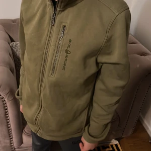 Olivgrön jacka från Sail Racing - Snygg olivgrön jacka från Sail Racing med dragkedja och huva. Jackan har en bröstficka med dragkedja och logotyp på bröstet. Insidan är fodrad med mjukt material för extra komfort. Perfekt för kyligare dagar. De går diskutera pris!