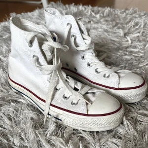 Vita Converse All Star high-top sneakers - Klassiska vita Converse All Star high-top sneakers med snörning och rund tå. Skorna har den ikoniska blå och röda randen på sulan samt Converse-loggan på sidan. Skorna är ett par år gamla, men har bara använts en gång🥰