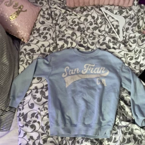 Ljusblå sweatshirt med San Fran-tryck från 157 - Ljusblå sweatshirt från 157 med vitt tryck där det står 'San Fran CA' på bröstet. Tröjan har rund halsringning och långa ärmar. Perfekt för en avslappnad och cool stil.