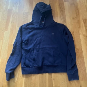 Mörkblå hoodie från GANT - Säljer en mörkblå hoodie från GANT med klassisk huva och magficka. Tröjan har diskret GANT-logga broderad på bröstet och är tillverkad i mjukt bomullsmaterial. Använd ett par gånger men inga tecken på användning! Skulle säga att den är i en stor S liten M