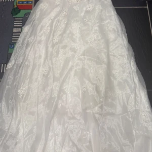 Vit långklänning med spets och pärlor - Säljer en vit bröllops  långklänning med vackra spetsdetaljer och pärlor. Klänningen har en elegant, utsvängd form och är dekorerad med blommiga broderier och glittrande pärlor. Under kjol, handskar igår, klänningen är minskad . Den var str M . Minskad till xs. Går o göra den större på bröst p midjan . Har bara sytt in den. Köpt för 8000 efter rea. 