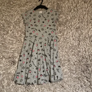 Grå kortärmad klänning med tryck - Grå klänning med kort ärm och svart samt rosa tryck med text och symboler som stjärnor, blixtar och läppar. Klänningen har rund halsringning och utställd kjol, perfekt för en avslappnad och lekfull stil.