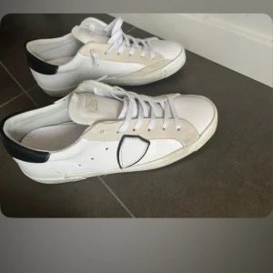 Vita sneakers från Philippe Model - Säljer ett par vita sneakers från Philippe Model med svarta detaljer på häl och sidan. Skorna har snörning och en platt sula. Klassisk och stilren design med inslag av beige mocka kring tån och snörningen. Dessa är i sprillans nytt skick använda ett fåtal gånger 