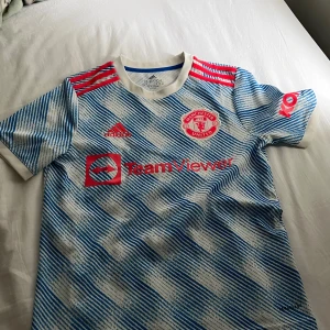 Manchester United bortatröja 2021/22 - Säljer en Manchester United bortatröja från Adidas med blått och vitt randigt mönster, röda detaljer och klubbmärke. Kortärmad modell med rund hals och TeamViewer sponsortryck på bröstet.