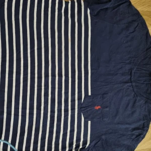 Randig t-shirt från Ralph Lauren - Mörkblå och vit randig t-shirt från Ralph Lauren med klassisk passform. T-shirten har korta ärmar, bröstficka med röd broderad logga och rund halsringning. Perfekt för en avslappnad stil.