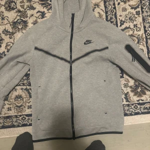 Grå zip-hoodie från Nike - Säljer en grå hoodie från Nike med hel dragkedja och svart logga på bröstet. Jackan har huva, svarta detaljer och en ficka med dragkedja på ärmen. Perfekt för en sportig och avslappnad stil. Storlek xs men passar S!