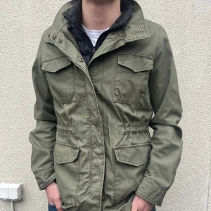 Olivgrön fieldjacka från Dobber - Säljer en olivgrön fieldjacket från Dobber med flera praktiska fickor och dragkedja. Jackan har en ståkrage och långa ärmar, perfekt för våren som kommer nu. Den har en stilren design med knappdetaljer vid kragen.