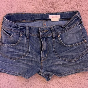 Blå jeansshorts med fickor - Klassiska blå jeansshorts med fem fickor, bälteshällor och knappgylf. Bakfickorna har lock med knapp. Perfekta för varma dagar och enkel att matcha med olika toppar.