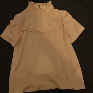 Ljusrosa blus med volangdetaljer - Söt ljusrosa blus med korta ärmar och volang längs bröstet och runt kragen. Blusen har volang och dragkedja i nacken.