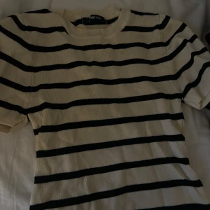 Randig stickad tröja från Ginatricot - Säljer en krämvit och svart randig stickad tröja från Ginatricot. Tröjan har korta ärmar och rund halsringning. Perfekt för en avslappnad och stilren look.