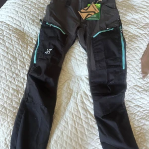 Svarta friluftsbyxor från RevolutionRace - Säljer ett par svarta friluftsbyxor från RevolutionRace med turkosa dragkedjor och elastisk midja. Byxorna har flera praktiska fickor och är perfekta för utomhusaktiviteter. Modellen är smal och har förstärkta partier på knän och bak.