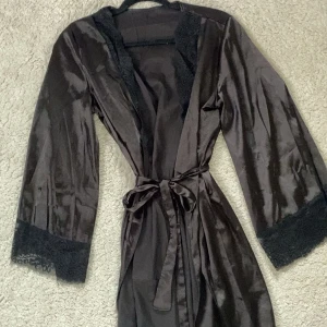 Svart satin morgonrock med spetsdetaljer - Säljer en svart morgonrock i satinmaterial med fina spetsdetaljer längs ärmslut och ”kragen”. Använd fåtal gånger💕💕💕Finns ingen lapp så vet inte storlek eller märke, men jag gissar på xs/s