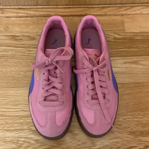 Rosa och lila sneakers från Puma - Säljer ett par snygga sneakers från Puma i rosa mocka med lila detaljer och gummisula. Skorna har klassisk snörning och guldfärgad Puma-logga på sidan. Perfekta för dig som vill sticka ut med färg på fötterna! Andvända inne bara cirka 5 gånger, pris kan diskuteras 