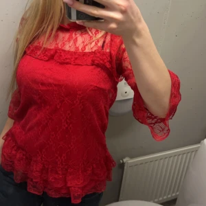 Röd spetsblus med volangdetaljer - Säljer en röd blus i spets med volangdetaljer och trekvartsärm. Blusen har en hög halsringning och är genombruten i materialet vilket ger en romantisk känsla. Perfekt för dig som vill sticka ut med färg och detaljer.