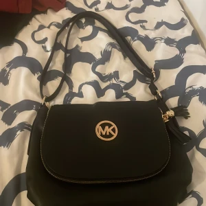 Svart axelväska från Michael Kors - Snygg svart axelväska från Michael Kors som inte säljs längre. Väskan har guldfärgade detaljer och dekorativ tofs och har ett stort fack, innerfickor och dragkedja både fram och bak. Perfekt för dig som vill ha en stilren och praktisk accessoar. Pris går att diskuteras.