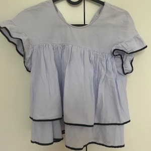 Ljusblå volangtopp med svarta kanter - Säljer en ljusblå topp med volanger och svarta kanter. Toppen har korta ärmar med volangdetaljer och en lös passform. Perfekt för vår och sommar!