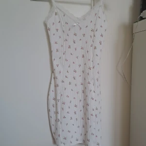 Vit blommig ribbad klänning med spetsdetaljer - Supersöt vit klänning med små rosa blommor, ribbad struktur och spets längs urringningen. Axelbandslös modell med liten rosett framtill. Perfekt för sommarens varma dagar! Figur sydd. Har en fläck på baksidan 🦄🦄🍾🍾🎉😝😝