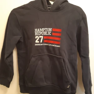 Mörkblå hoodie från Hampton Republic - Mörkblå hoodie från Hampton Republic med tryck och broderi framtill. Tröjan har känguruficka och huva, perfekt för en avslappnad stil. Klassisk design med röda och vita detaljer på bröstet.
