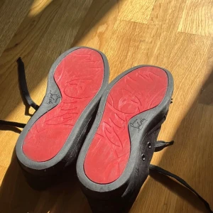 Svarta sneakers med nitar och röd sula - Säljer ett par svarta sneakers från Christian Louboutin med ikonisk röd sula och svarta nitar på tån. Skorna har snörning och är tillverkade i mocka med detaljer i skinn. Perfekta för dig som vill sticka ut med din stil. Dustbags tillkommer
