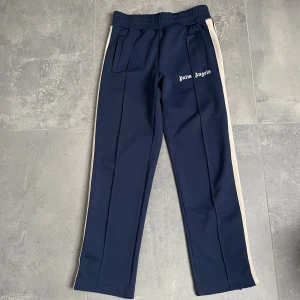 Mörkblå Palm Angels byxor - Säljer ett par mörkblå Palm Angels byxor med vita revärer längs sidorna och logga tryckt på vänster lår. Byxorna har resår i midjan och raka ben för en avslappnad look. Äkthetsbevis finns! 