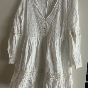 Vit långärmad klänning med spetsdetaljer från ZARA - Säljer en romantisk vit klänning med lång ärm och v-ringning. Klänningen har fina spetsdetaljer vid ärmslut och midja samt dekorativa knappar framtill. Perfekt för sommarens alla tillfällen!