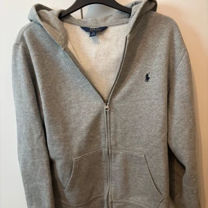 Grå hoodie med dragkedja från Polo Ralph Lauren - Säljer en klassisk grå hoodie från Polo Ralph Lauren med dragkedja och huva. Tröjan har mjukt foder på insidan och den ikoniska broderade loggan på bröstet. Perfekt för en avslappnad stil.