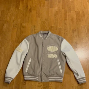 Grå och vit varsityjacka från Zara - Säljer en snygg varsityjacka från Zara i grått och vitt med broderade detaljer och tryck. Jackan har ribbade muddar, knappar framtill och två fickor. Perfekt för dig som gillar streetwear och vill sticka ut med stilrena detaljer.