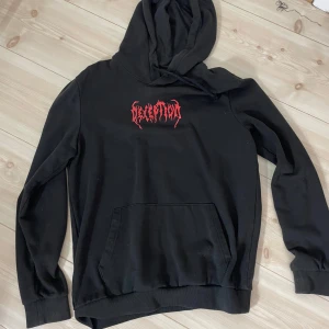 Svart hoodie med röd DECEPTION-tryck från Zalando  - Svart oversized hoodie med rött tryck 'DECEPTION' på bröstet och stor grafik på ryggen. Huva och magficka framtill. Perfekt för dig som gillar streetwear och statement-plagg.