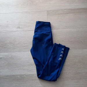 Blå träningstights med hög midja - Säljer ett par blå träningstights med hög midja och diskret mönster på ena benet. Tightsen har fickor på sidorna och är i stretchigt material som passar perfekt för träning.