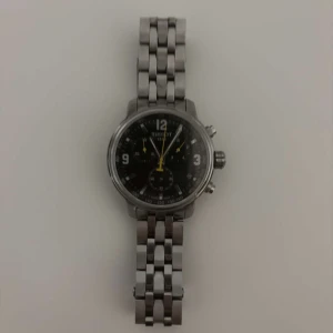 Tissot kronograf herrklocka i silver - Säljer en stilren Tissot herrklocka med kronograf och svart urtavla. Klockan har silverfärgat armband i metall och tydliga siffror samt gula detaljer på visarna. Perfekt accessoar för dig som gillar klassisk design.