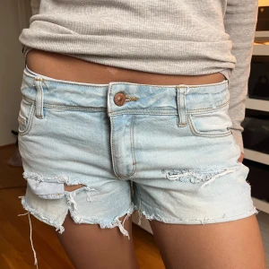 Ljusblå lågmidjade jeansshorts med slitningar - Säljer ett par ljusblå jeansshorts med råa kanter och slitna detaljer. Klassisk femficksmodell med knapp och dragkedja framtill. Perfekta för varma dagar och en avslappnad stil. Jeansshortsen är låg midjade. Skriv för mått osv. Priset går att diskutera. 