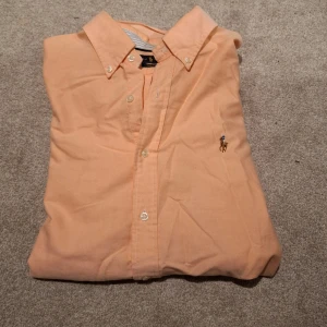 Persikofärgad skjorta från Ralph Lauren - Säljer en klassisk persikofärgad skjorta från Ralph Lauren med button-down krage och broderad logga på bröstet. Skjortan har långa ärmar och knäppning framtill. Perfekt för en stilren look!