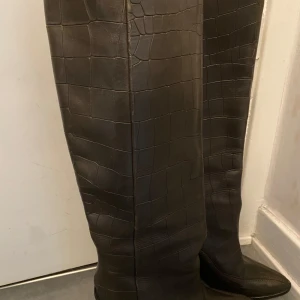 Bruna höga stövlar med krokodilmönster - Säljer ett par bruna höga stövlar med krokodilmönster och spetsig tå. De är i ny skick knappt använda de är i äkta läder i storlek 36