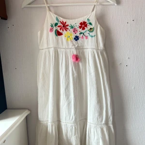 Vit sommarklänning med blommor  - Superfin vit klänning från H&M med smala axelband och färgglada broderade blommor framtill. Lätt och luftig modell med volang nertill och en liten rosa tofs. Perfekt nu till sommaren! Otroligt bra skick och tyget är tjockt och fint. Storlek 140 💘